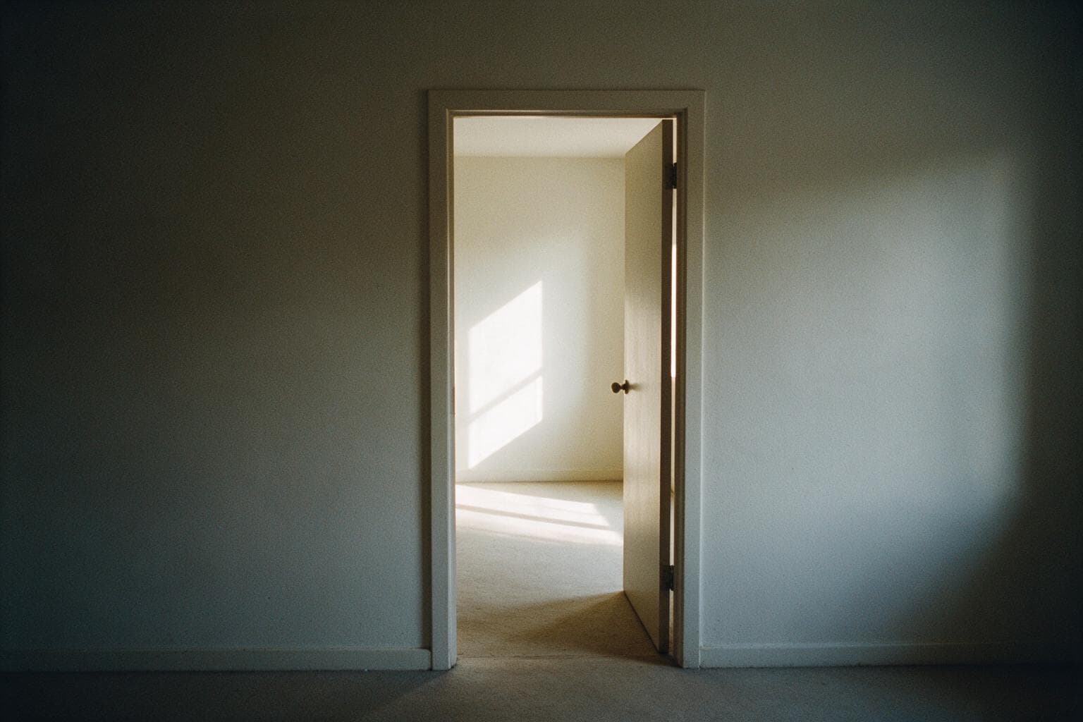 An open doorway in a plain interior wall leading to a sunlit room