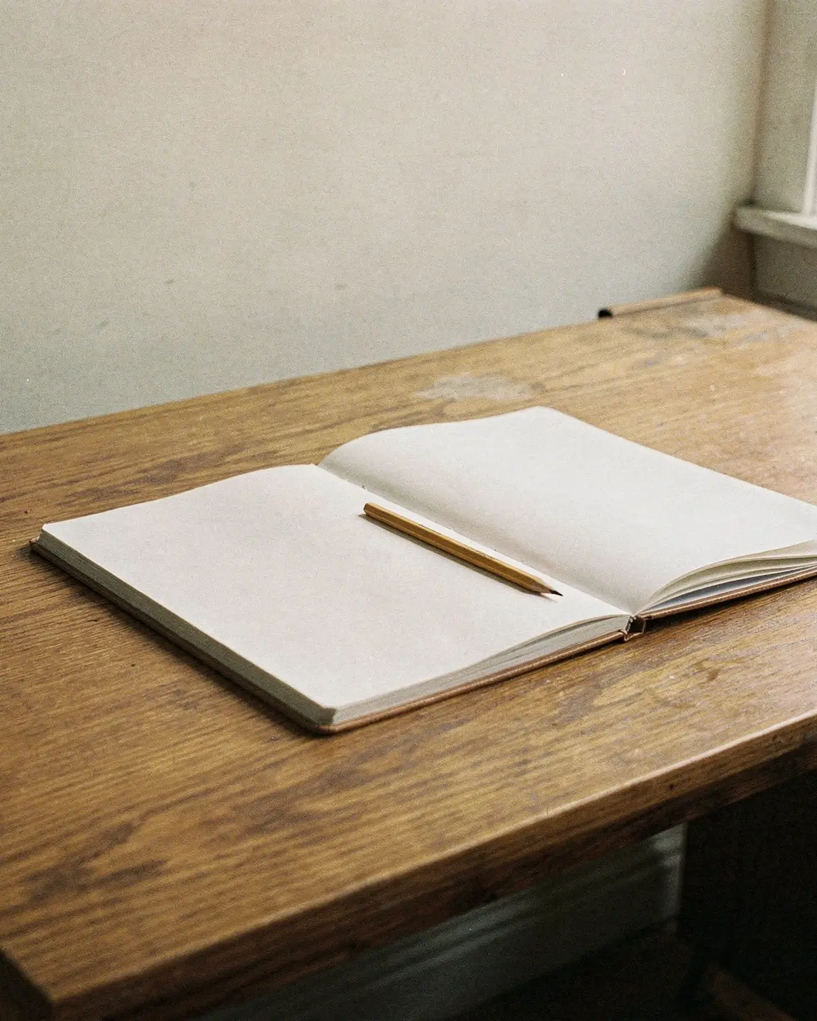 An open paper notebook and a plain pencil on an oak desk in natural daylight